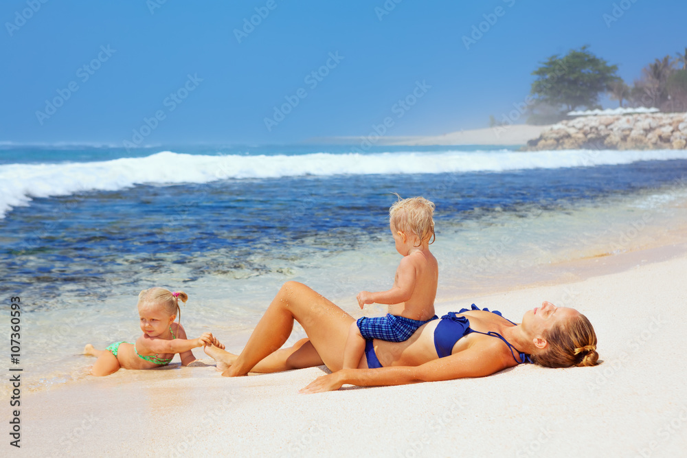 Happy family - mother, baby son and daughter lying on sand beach and look at sea surf after swimming in clear water. Active parents and people outdoor activity on tropical summer vacations with child