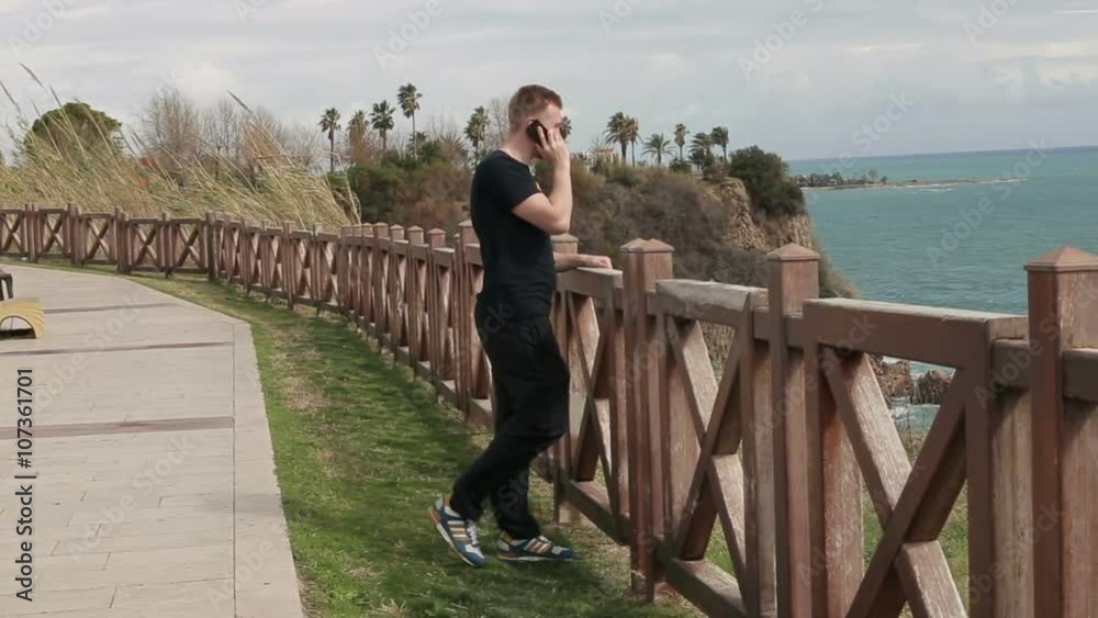 Young happy man talking using a smartphone while standing on the waterfront looking at the sea Young happy man talking using a smartphone while standing on the waterfront looking at the sea