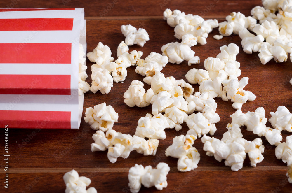 Red white striped container box lying tipped over on wooden surface with popcorn spread out Red white striped container box lying tipped over on wooden surface with popcorn spread out