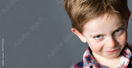 closeup portrait of a young mischievous child with freckles teasing and grumbling with fun look and joke, copy space on grey background studio photo