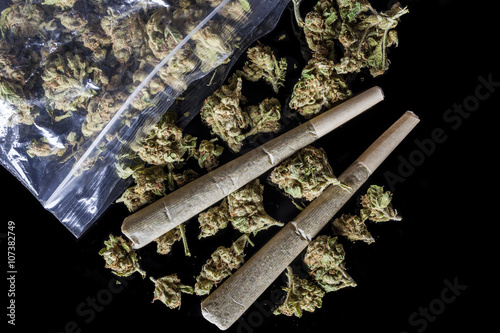 Pile of medical cannabis dried buds scattered from nylon package and two marijuana joints on black background directly from above