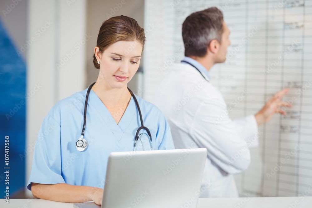 Female doctor working on laptop with colleague pointing at chart in hospital Female doctor working on laptop with colleague pointing at chart in hospital