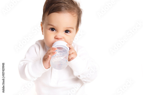 Portrait of a little boy,brunette with brown eyes,dressed in a white shirt and a white diaper,barefoot posing in Studio,sitting on a white background with a bottle for baby food,drinking water