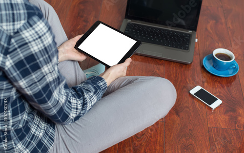 The woman was wearing a blue plaid shirt with a tablet cross legged sitting on the red wood floor, floor placed a phone, a cup of coffee, a notebook and a pen and a laptop computer.