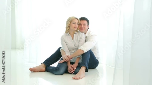 Husband and wife sitting on the floor next to each other near a large window, they hug, looking at the camera.Happy family in white shirts and jeans. Man and woman smiling, hugging. photo