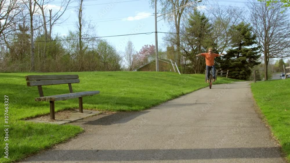 Young man balances on unicycle, then rides past camera, smiling as he passes in a suburban park on a sunny spring day. Recorded hand-held with a wide lens in slow motion 4K at 60fps. Young man balances on unicycle, then rides past camera, smiling as he passes in a suburban park on a sunny spring day. Recorded hand-held with a wide lens in slow motion 4K at 60fps.