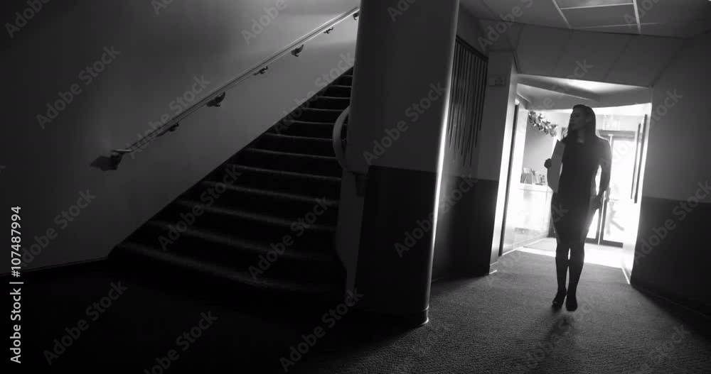 Woman walks through noir-style theater lobby, past staircase.  Silhouetted man in a hat follows her, then goes up stairs casting a dark shadow.  Wide shot, black and white, originally recorded in 4K.