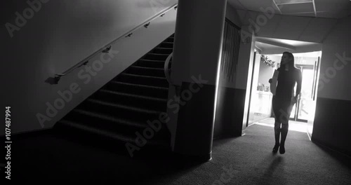 Woman walks through noir-style theater lobby, past staircase.  Silhouetted man in a hat follows her, then goes up stairs casting a dark shadow.  Wide shot, black and white, originally recorded in 4K.