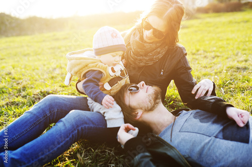 A young and happy family is resting with his young son in the park in the summer. Parents smiling. Sunglasses. Leather jackets. Sunset. The sun. photo