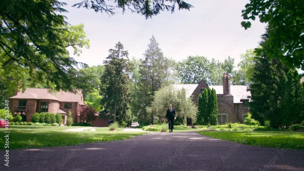 Man in suit walks up driveway, towards camera, in wealthy neighborhood in the US Mid West. Locked off camera, low viewpoint. Originally recorded in 4K. Man in suit walks up driveway, towards camera, in wealthy neighborhood in the US Mid West. Locked off camera, low viewpoint. Originally recorded in 4K.