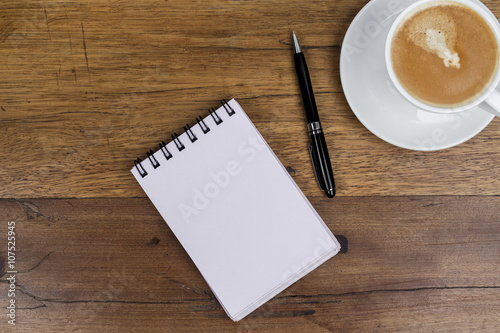 White sheets notebook with spiral in the center of the wooden dark brown table with black pen aside and cup of coffee on top right from above