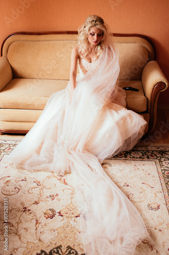 Gorgeous blonde bride in stylish wedding dress posing on sofa