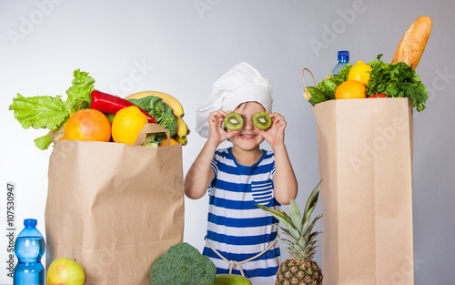 Little happy girl in chef hat with big packages of products and sliced kiwi near eyes photo