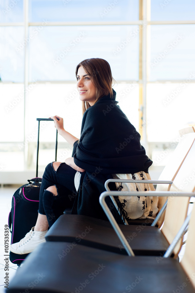 Fototapeta premium Airport Young female passenger at gate waiting in terminal while waiting for her flight. Air travel concept with young casual woman sitting with hand luggage suitcase.