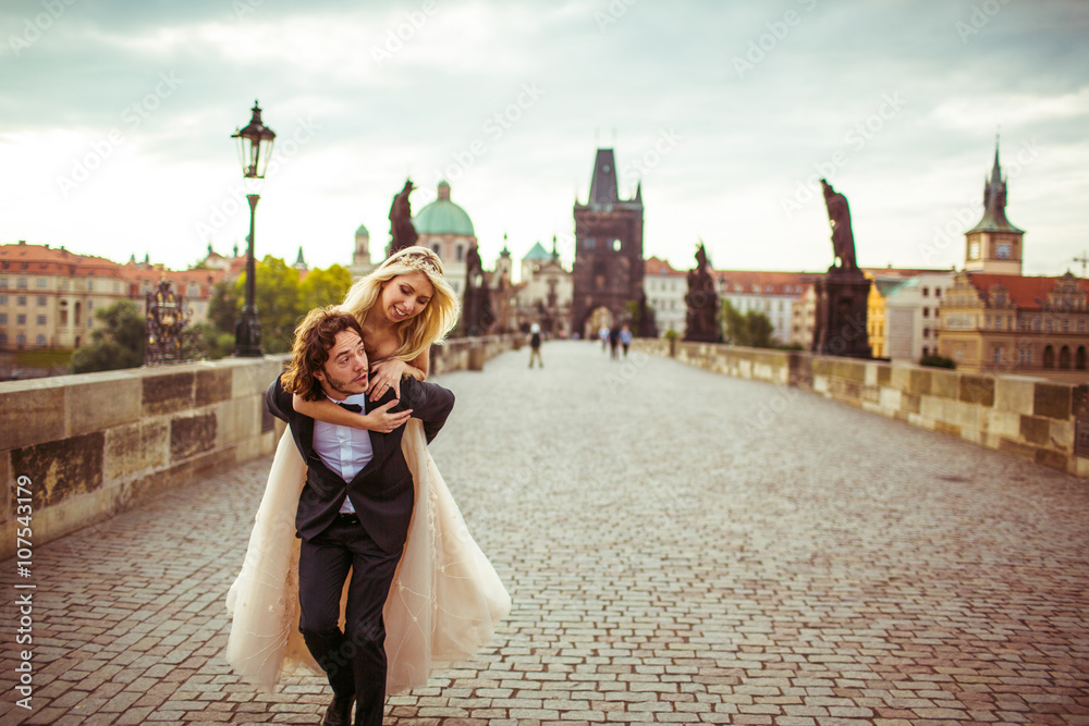 Fototapeta premium Romantic handsome groom carrying happy beautiful bride on bridge