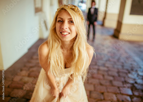 Beautiful blonde bride posing near buildng, groom in background photo