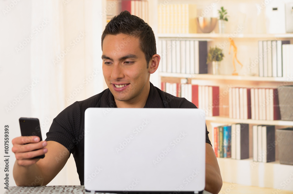 Young hispanic man sitting by desk working on white laptop and looking at mobile phone Young hispanic man sitting by desk working on white laptop and looking at mobile phone