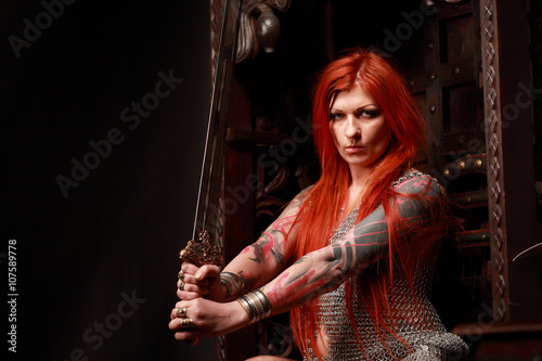 Studio shot of beautiful young redhead girl with multiple tattoo in chainded armour with the sword