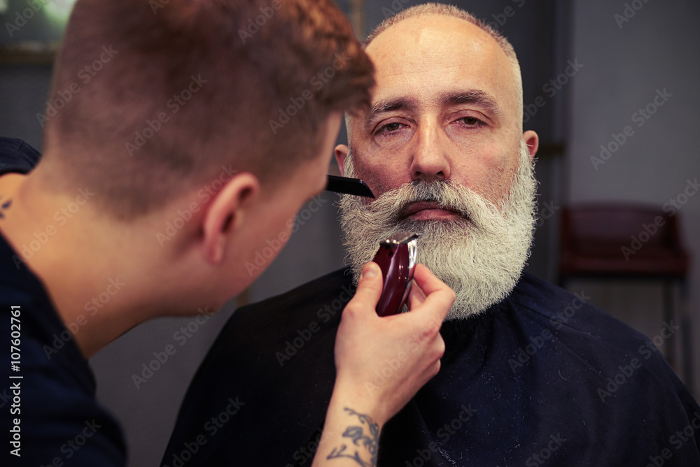 Fototapeta premium Barber making haircut to handsome attractive senior man with bea