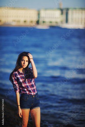 beautiful brunette girl in shorts and a plaid shirt straightens the hair on background of river embankment © raisondtre