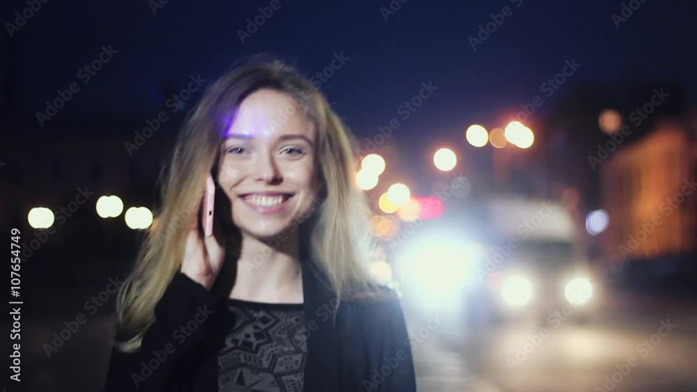 Beautiful and Cute Girl Talking on the Phone in Evening, Standing in the Night City. in the Background, Street Lamps and Lights. Drive Cars. Beautiful and Cute Girl Talking on the Phone in Evening, Standing in the Night City. in the Background, Street Lamps and Lights. Drive Cars.