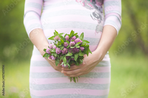 Closeup shot of pregnant woman with pink clover bouquet on her belly dressed in striped pink and grey dress
