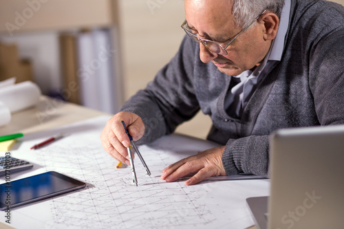 Senior architect working on construction blueprint in office, he draws with a divider.