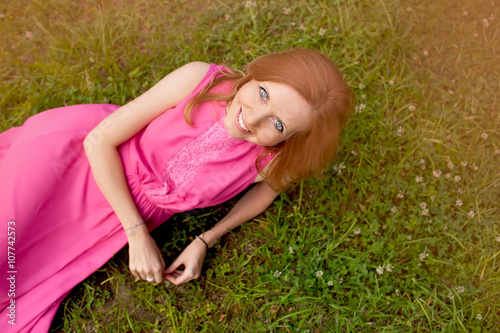 The young girl in a pink dress lies on a grass in park and looks in a camera. Top view.