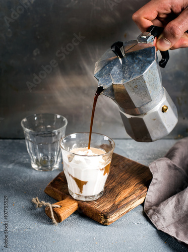 Glass of coffee with ice cream on rustic wooden board. Drink is poured from steel Italian Moka pot held by man's hand photo