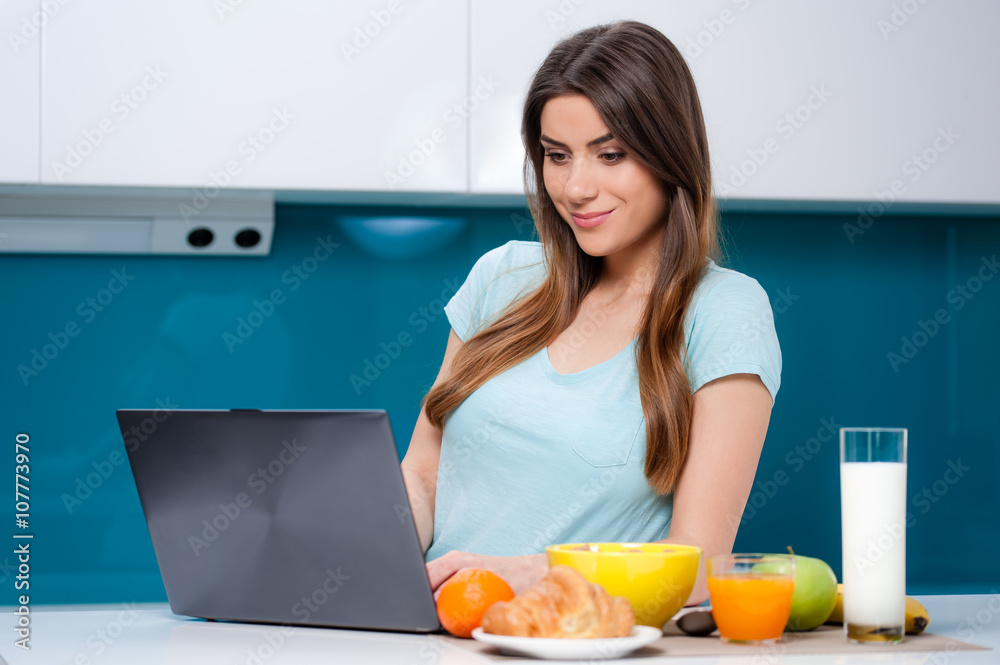 Casual woman eating breakfast and working or catching up on her social media on her laptop as she sits at the dining table at home