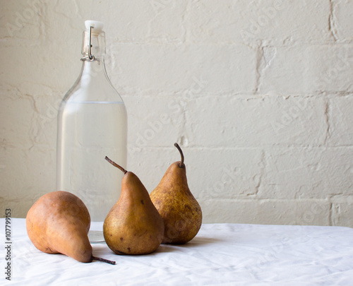 Three beurre bosc pears and a glass bottle of water on a white tablecloth against a white brick wall