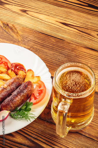 Mug of beer and grilled sausages with fried potatoes, sliced tomatoes, fresh produce, ketchup in plate on wooden table