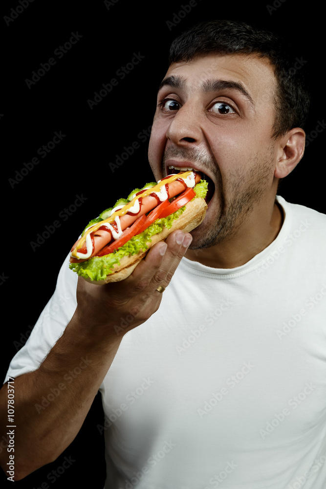 Young armenian man eating a messy hotdog isolated on black background Young armenian man eating a messy hotdog isolated on black background