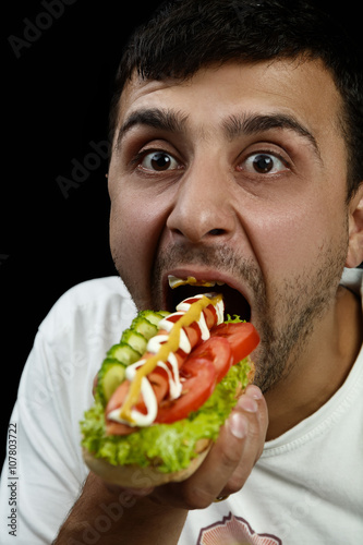 Young armenian man eating a messy hotdog isolated on black background