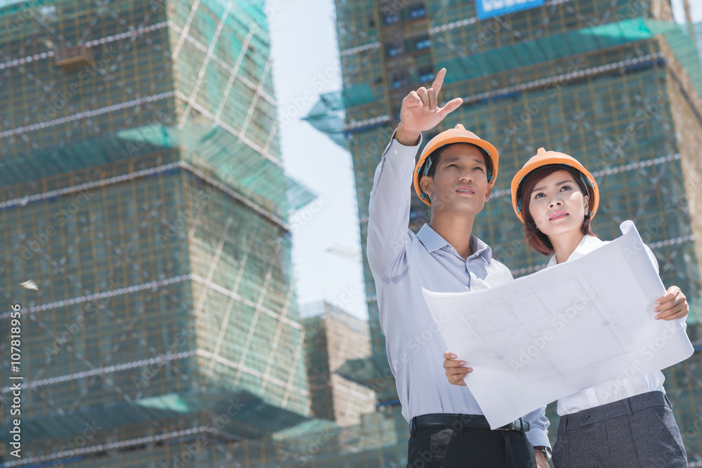 Young Vietnamese engineers comparing blueprint with actual building Young Vietnamese engineers comparing blueprint with actual building