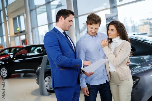 Salesman talking to a young couple at the dealership showroom. © Nejron Photo