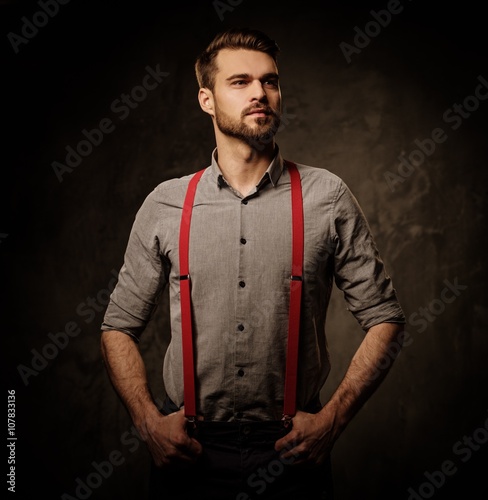 Young handsome man with beard wearing suspenders and posing on dark background.