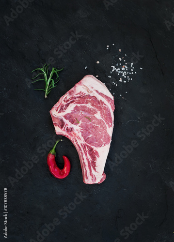 Raw fresh meat ribeye steak with salt, chili pepper and rosemary over black slate stone background, top view