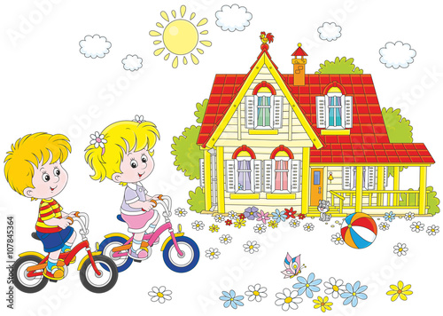 A little girl and a little boy riding bikes on a sunny summer day near a country house