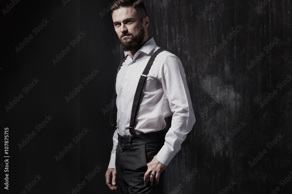 Attractive bearded man in a white shirt and suspenders standing near dark wall. He is strong, courageous and serious. Dark room, night and harsh shadows.