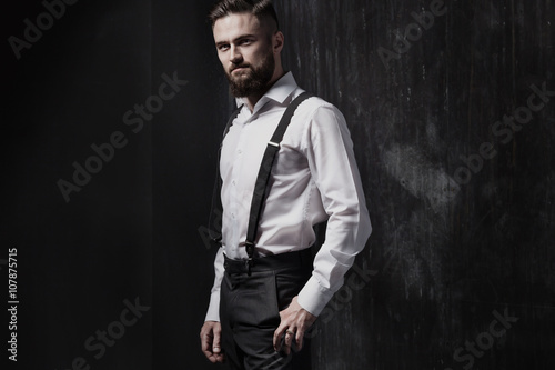 Foto Attractive bearded man in a white shirt and suspenders standing near dark wall