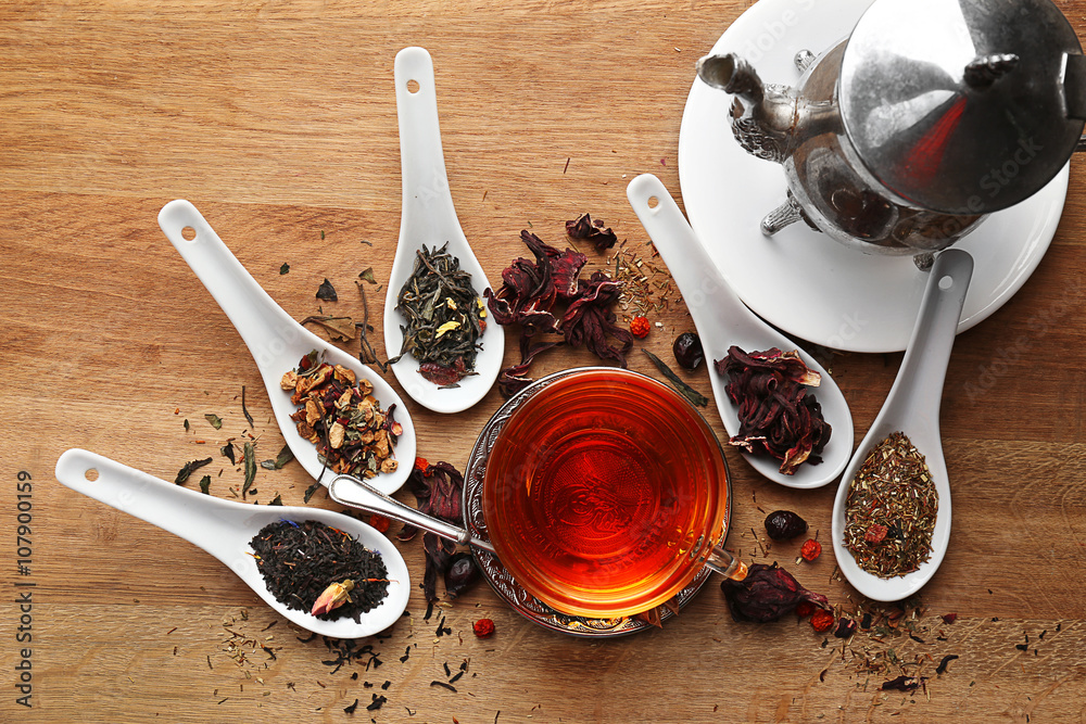 Fototapeta premium Tea concept. Different kinds of dry tea in spoons. Glass cup of tea on wooden background