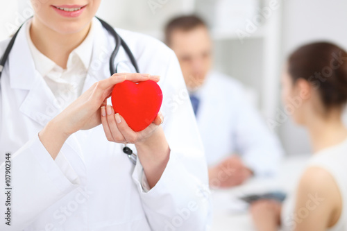 Female doctor with stethoscope holding heart.  Doctor and patient sitting in the background
