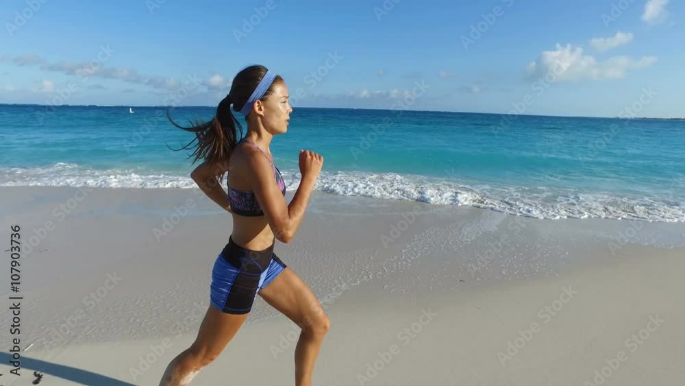 Woman jogging and running on beach. Fit young female runner in sportswear is training on idyllic beach. Runner is exercising in nature on sunny day. ACTION CAMERA TRACKING SHOT.