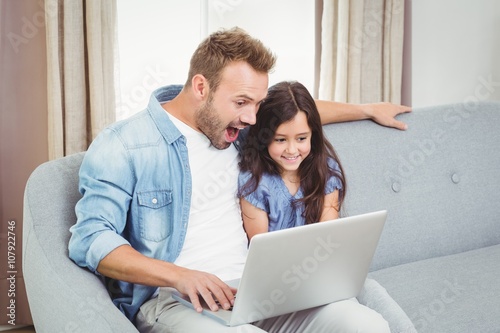 Surprised father and daughter using laptop while sitting on sofa at home