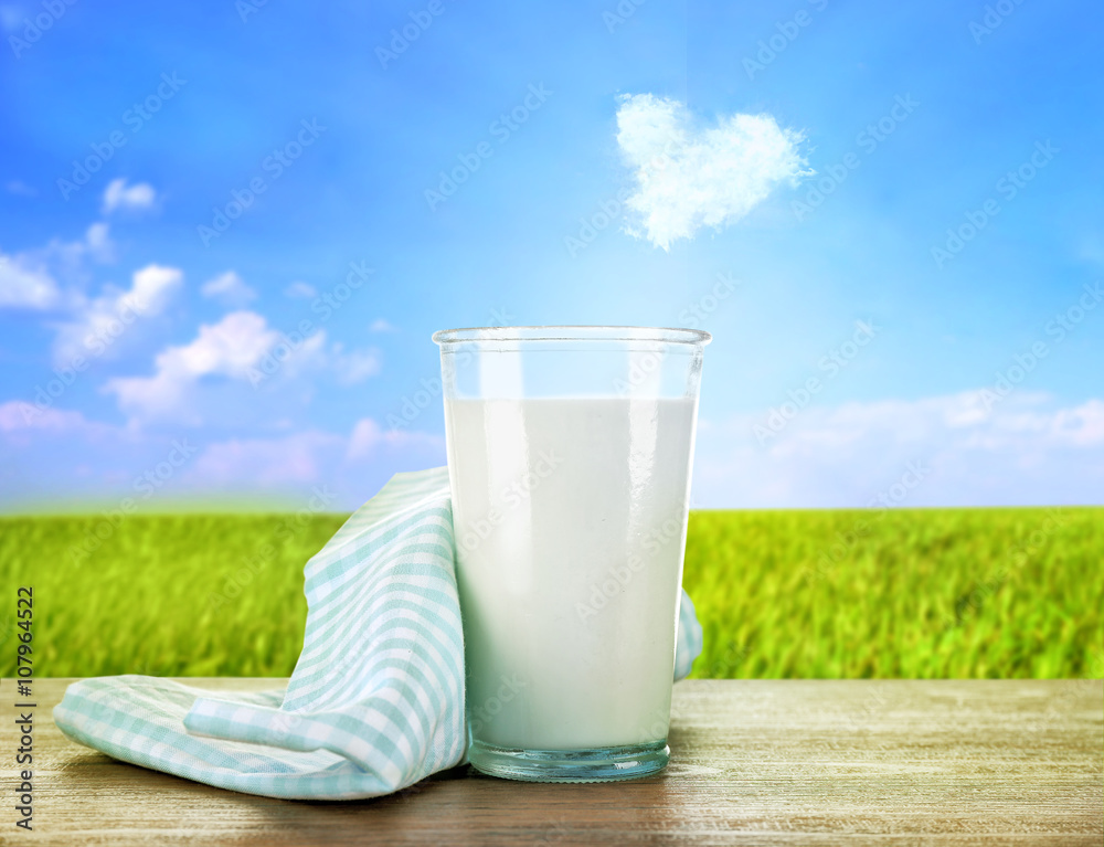 Glass of milk on wooden table against green field and blue sky background Glass of milk on wooden table against green field and blue sky background