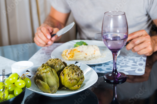 Classy dinner table setting, cooked artichokes sitting in middle, mans arms visible holding cutlery next to white plate of crepe serving