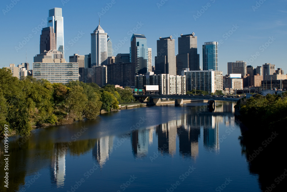 The skyline of Philidelphia. The Schuylkill river is in the foreground. The tallest buildings are on the left side. The buildings are reflected in the surface of the river. 
