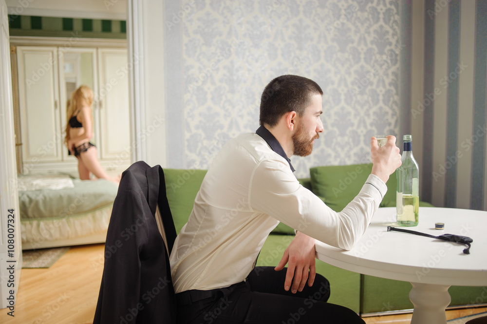 Portrait of young concerned man in suit sitting at the table with shot of alcohol in his hand and beautiful sexy lady in lingerie on the background. Relationship concept. Portrait of young concerned man in suit sitting at the table with shot of alcohol in his hand and beautiful sexy lady in lingerie on the background. Relationship concept.