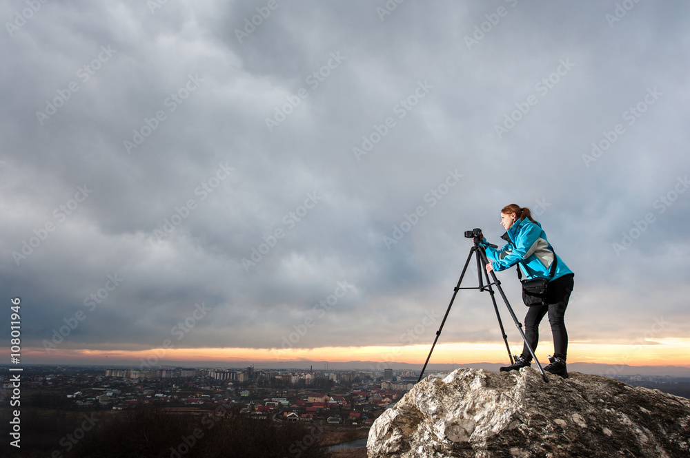 Young female photographer is standing with her camera on tripod on the big rock at city overview point. Woman is taking picture of the cityscape in the evening